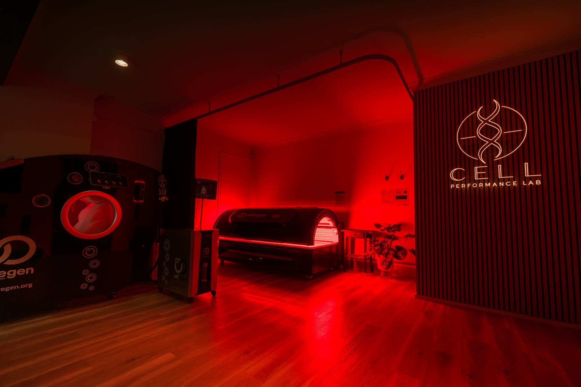 The treatment room at CELL Performance Lab in red light, with the hyperbaric chamber, red light bed and CELL logo wall